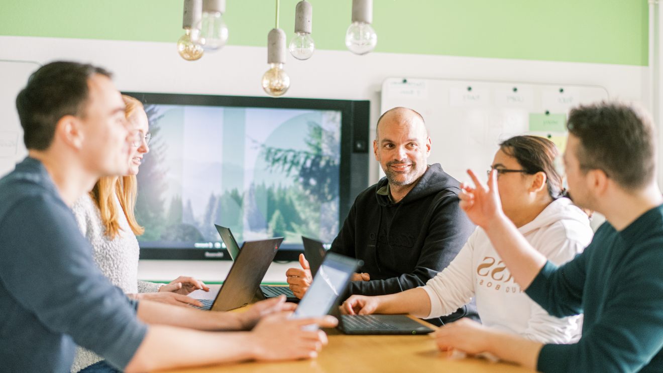 Fünf Personen diskutieren an einem Tisch vor einem Bildschirm